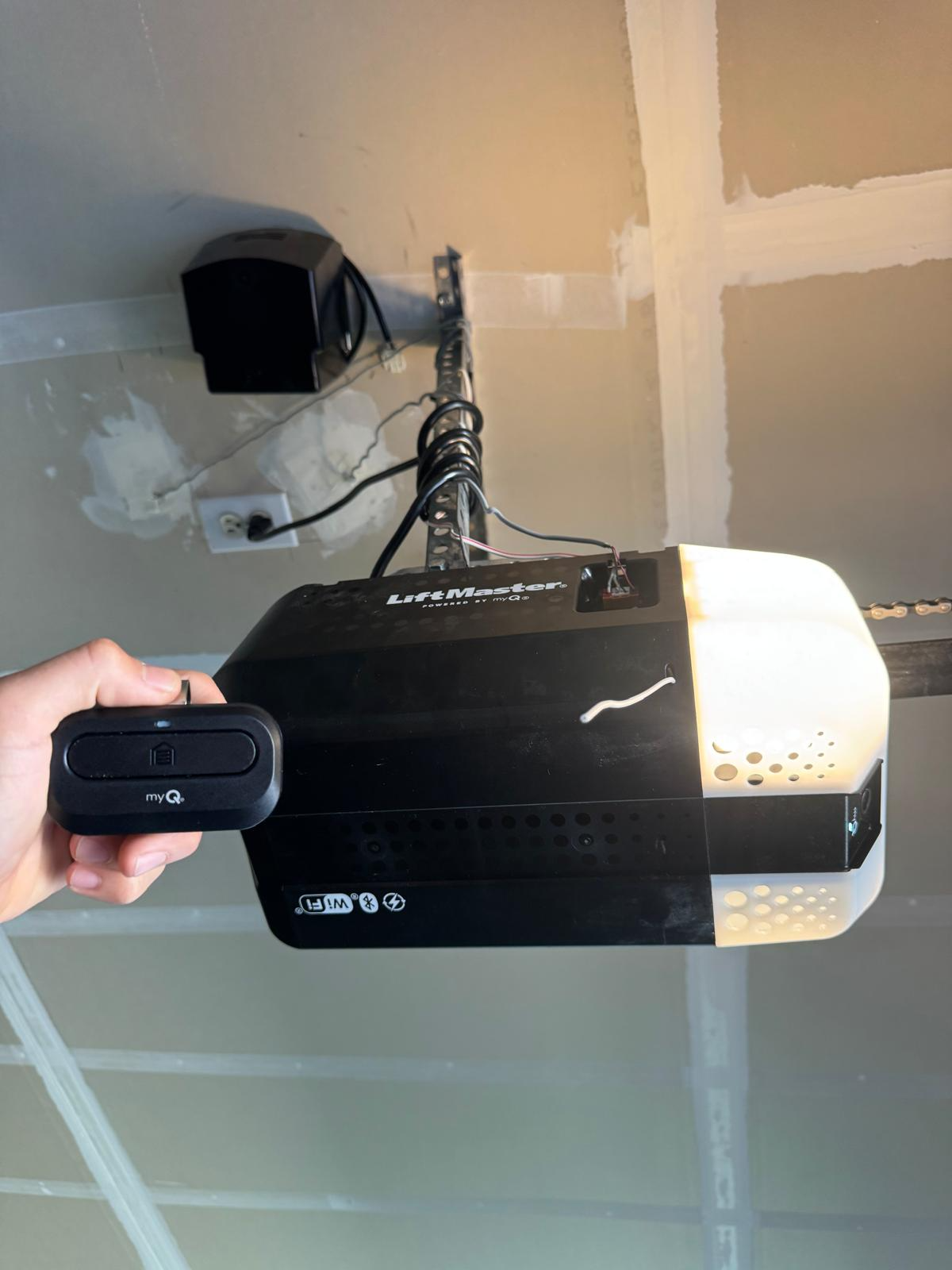LiftMaster garage door opener with its light lit on a drywall ceiling, with a hand holding a myQ remote control
