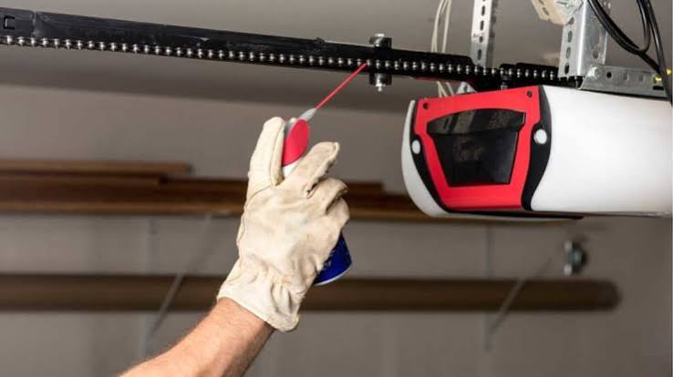 Technician spraying lubricant on a garage door opener drive chain during a maintenance service