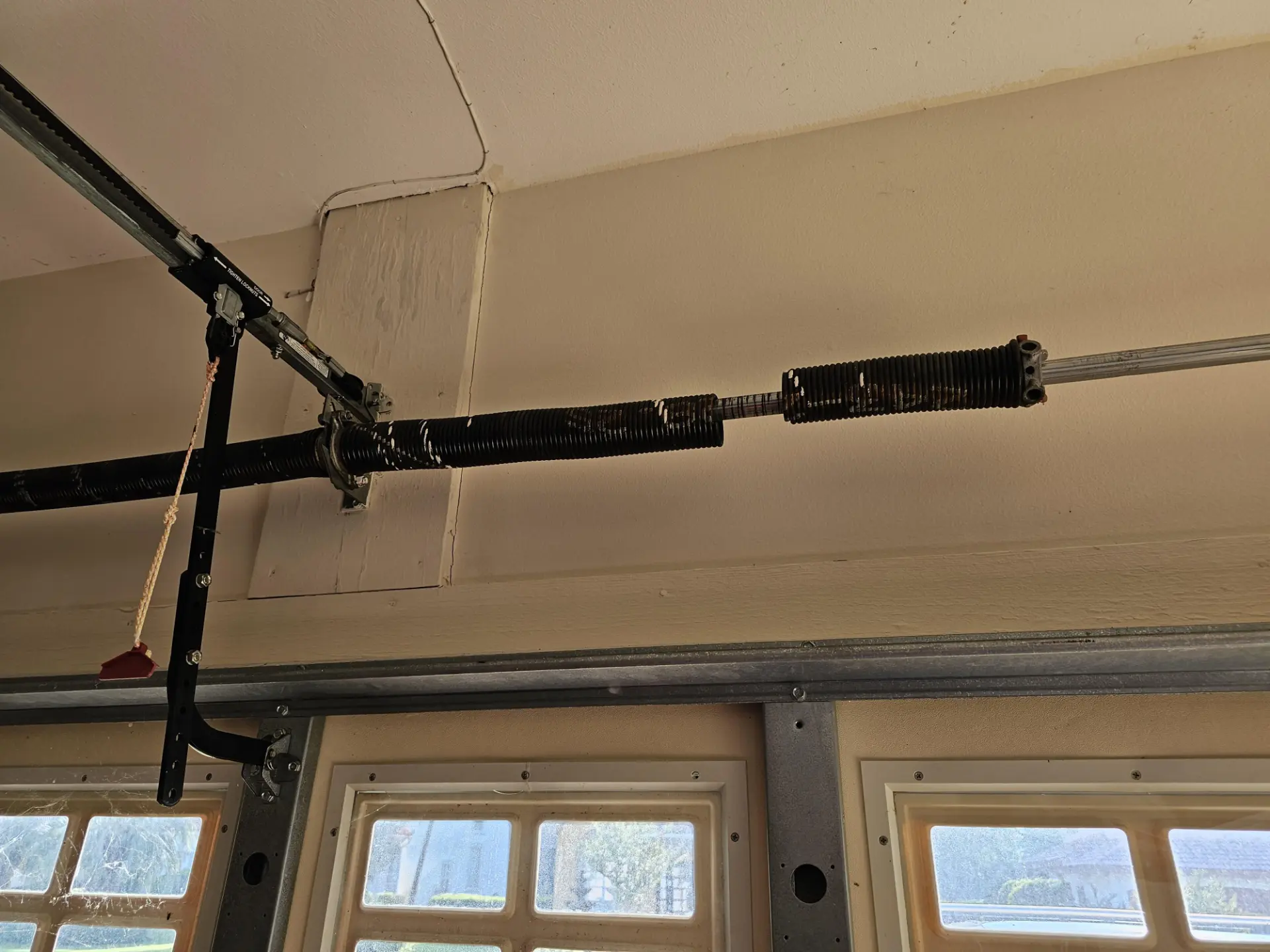 Broken black torsion garage door spring snapped in two on the shaft above a windowed garage door, with the opener arm and red emergency release cord visible