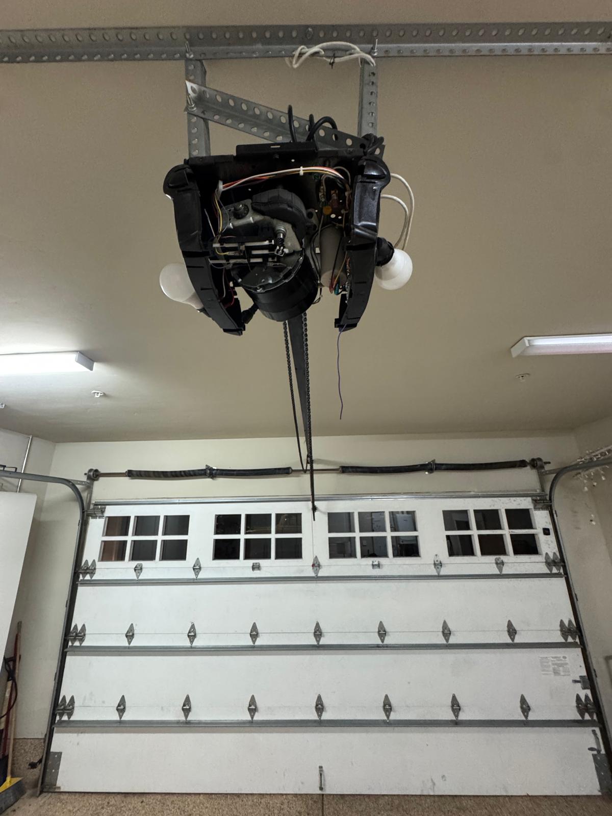 Garage door opener motor unit with its cover removed, exposing the motor, drive chain, wiring and light bulbs above a closed white garage door